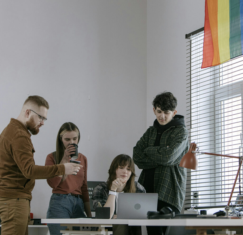 Grupo de jóvenes con bandera LGBTIQ+ alrededor de computadora. Sección Nosotros